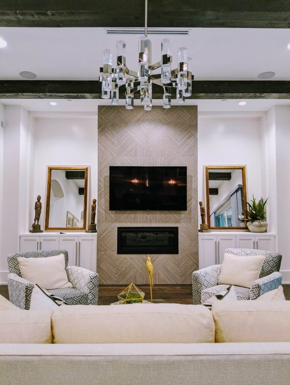 Bellaire Estate — The living room centers on a herringbone-wood fireplace wall and crystal chandelier, flanked by lit display niches and patterned swivel chairs.