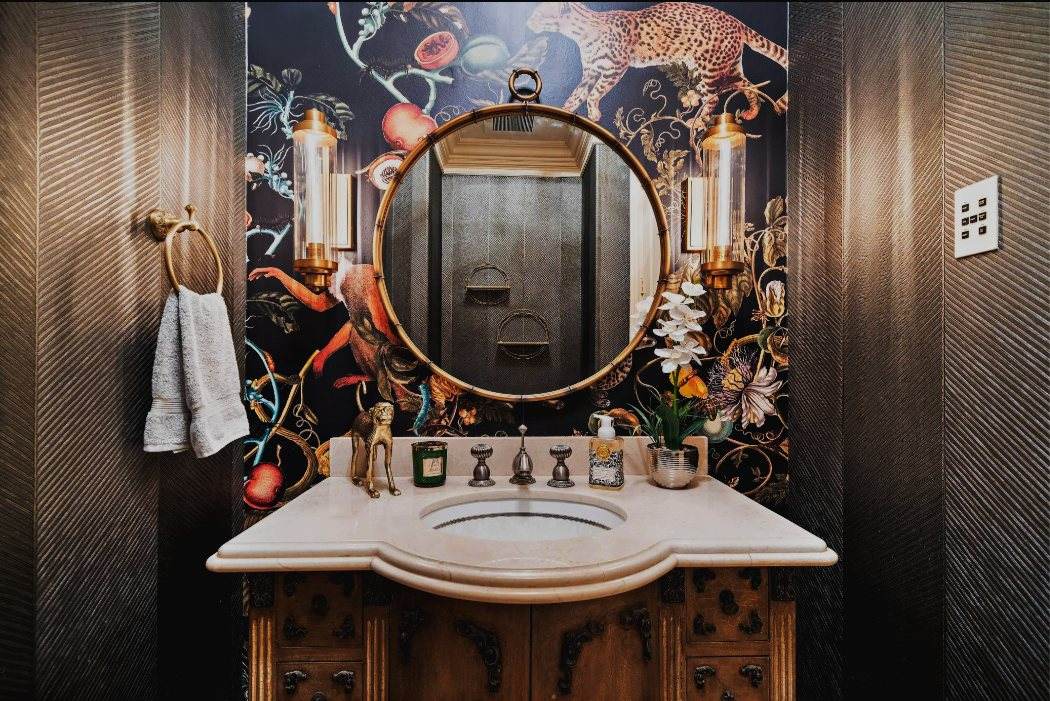 Lakes of Parkway Residence — A dramatic powder room — botanical wallpaper, herringbone wrap walls, an antique carved vanity, and a round brass mirror.