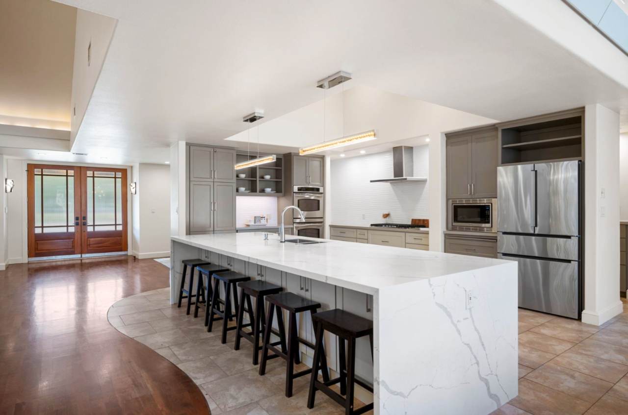 Memorial Residence — The renovated kitchen centers on a waterfall marble island with seating for five, framed by soft gray cabinetry and a linear pendant.